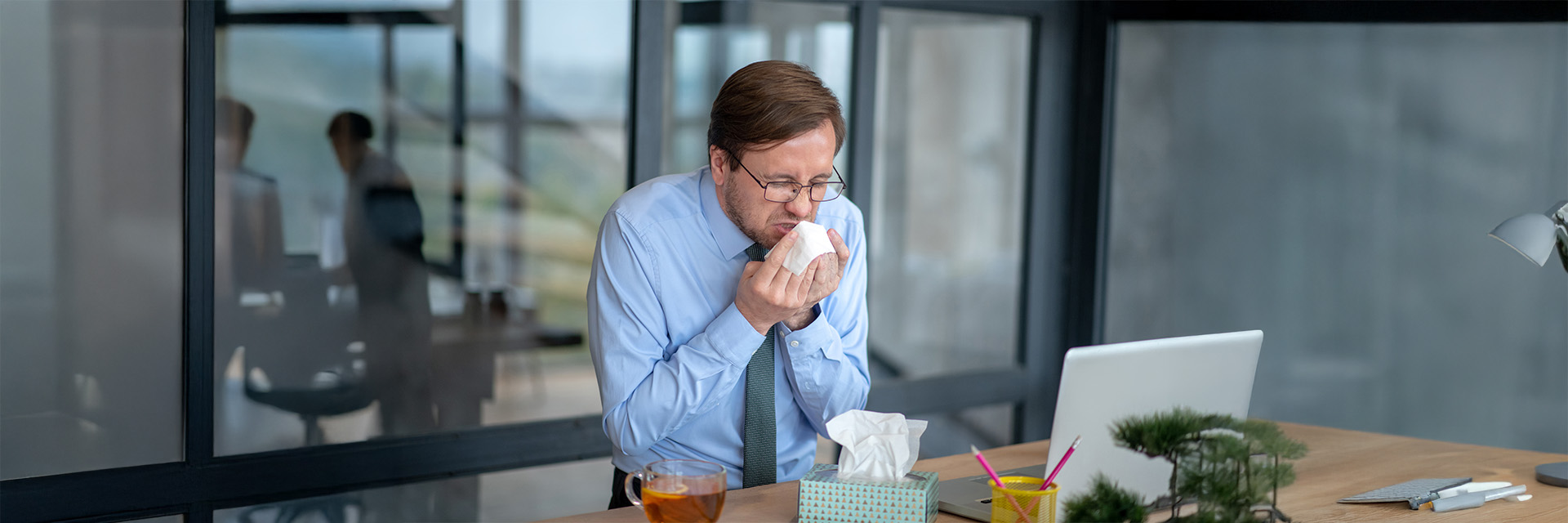 An office worker sneezing into a tissue while working at his desk.